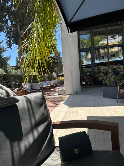 An outdoor seating area of a cafe in Paphos Municipality, Cyprus, is depicted. The foreground is dominated by a dark grey upholstered chair with a patterned cushion. A black quilted handbag with a gold-tone logo and chain strap rests on the seat of the chair. Sunlight casts shadows across the chair and the light-colored tiled floor.
In the midground, several rows of outdoor seating with woven chairs and tables are visible. A person in a white t-shirt is seated at one of the tables, holding a phone. Palm fronds hang from the left side of the frame, partially obscuring the view.
The background features a building with large windows reflecting palm trees and a blue sky. A concrete pillar is prominent on the left side of the building. Beyond the cafe, trees and other vegetation are present. The overall impression is of a bright, sunny day at a cafe.