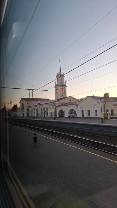 The image shows a train station in Volkhov, Russia, likely during sunrise or sunset. The station building is a light yellow, multi-story structure with arched windows and a prominent clock tower. The clock faces are illuminated, suggesting it is either early morning or late evening. Train tracks run alongside a brick-paved platform, and overhead wires are visible, indicating an electrified railway. Reflections in what appears to be a train window or glass structure show the building and the sky. The sky is a pale, hazy orange and blue, typical of twilight hours. No people are clearly visible, but a small dark object, possibly a piece of equipment or signage, is on the platform. The scene evokes a sense of quiet travel or anticipation. There is no visible text in the image.