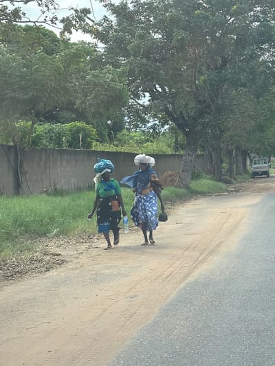 Two women are walking along the side of a road in Pemba, Mozambique. The woman in front is wearing a white headwrap, a blue patterned dress, and flip-flops. She is carrying a black object on her head and a water bottle in her hand. The woman behind her is wearing a blue headwrap, a green shirt, black shorts, and flip-flops. She is also carrying an object on her head. The road is paved on one side and dirt on the other. There is grass and a wall on one side of the road and trees on the other. A white truck is visible in the distance.