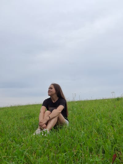 A young woman with long, dark brown hair sits in a lush green grassy field under a cloudy sky. She is wearing a black t-shirt and white sneakers. Her legs are crossed, and her arms are resting on her knees. She is looking off to her right, with a slight smile on her face. The background is a vast expanse of green grass and a pale, overcast sky. There are no visible buildings or structures in the immediate vicinity. The image appears to be taken outdoors in a rural or park-like setting. The location in the city of Гаревая, Russia is not discernible from the image itself.