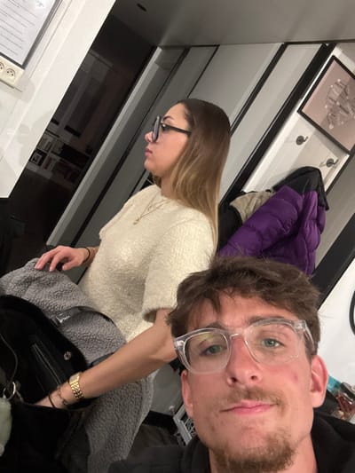 A selfie-style shot shows a man and a woman. The man, positioned closer to the camera, wears clear-framed glasses and has a beard. The woman, wearing glasses and a white sweater, is seen in profile. A purple jacket hangs in the background. The scene is indoors, with a picture frame visible on the wall. The lighting is soft, creating a casual atmosphere.