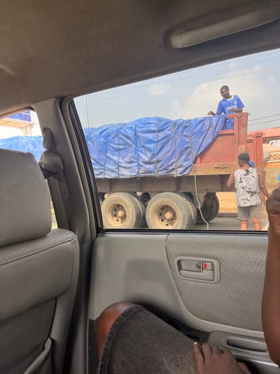 The image shows two people near a large truck loaded with cargo covered by a blue tarp. The scene is outdoors, likely in Nigeria, given the location context. One person, wearing a blue shirt with "QATAR" visible on it, is standing on the truck, appearing to be securing the tarp. Another person, wearing a patterned shirt and shorts, is also on the truck, facing away from the camera.
The truck has several large tires visible. The image is taken from inside a vehicle, with a car door and interior visible in the foreground. A hand is visible resting on the car door. The sky is overcast, suggesting it might be daytime, but the weather is not definitively clear. There are no strong emotional cues, but the activity suggests a practical task of securing cargo. The overall setting appears to be a roadside or a loading area.