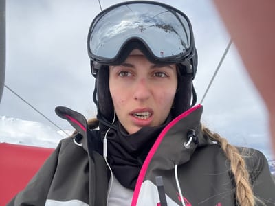 A selfie-style portrait captured on a ski lift shows a young woman, rotated for proper viewing. She is depicted from the upper chest upwards, with light brown or blonde hair styled in a braid that falls over her left shoulder. Her eyes are open, looking directly at the camera, and her mouth is slightly agape, revealing her teeth, with a neutral to light expression. She has some visible blemishes on her skin.
She is dressed in winter sportswear, including a dark olive green or grey ski jacket with white sections and bright pink trim on the zippers and edges. Beneath this, a black hooded layer and a white base layer are visible. White wired earphones are in her ears, with the wire draped over her jacket. A black ski helmet with fleece ear pads covers her head, and dark-framed ski goggles with reflective lenses are pulled up onto her forehead, obscuring her eyebrows. The goggles distinctly reflect a snowy mountain landscape and parts of a ski lift system, including cables or pylons.
The background shows an overcast sky and snow-covered terrain, characteristic of a high-altitude winter environment. To her right, a bright red component, likely part of the ski lift chair, is visible. A thin white rope or cable extends upwards on her left side, possibly part of the lift's structure or a safety element. The scene suggests a casual moment during a skiing trip in Val-d'Isère, France. No discernible text is visible on her clothing or equipment.