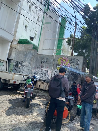 The scene takes place on a narrow street in Cimahi, Indonesia, located at the coordinates -6.884 latitude and 107.553 longitude. Two men stand on the pavement, one with a backpack and the other wearing a dark hoodie and blue jeans. The man with the backpack is facing the right, while the man in the hoodie is facing left, engaging in a conversation. Behind them, another man sits on a motorcycle, and a woman in a red shirt is standing next to the motorcycle. Several motorcycles are parked on the side of the street, and a large truck is visible in the background. The street is covered in a layer of cracked pavement, and there are several power lines overhead. Graffiti decorates a wall in the background. The wall features a mural of a cartoon face.