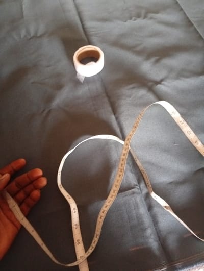 An overhead shot shows a hand on the left side of the frame, with fingers spread open and pointing downward. Beside the hand, a white measuring tape is draped across a dark grey fabric. The measuring tape curves upwards and to the right, with black numerical markings visible. Further to the right, a small roll of white tape sits on the fabric. The fabric appears to be slightly wrinkled, with folds and shadows creating texture. The scene is illuminated from above, casting soft shadows on the fabric. The location is the city of Gulak, Nigeria.