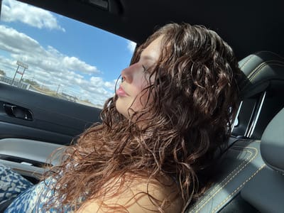 The image depicts a woman with wet, curly, brown hair, seen from a low angle inside a car. Her face is tilted upward, showing her lips and nose. The car's interior features a black dashboard with yellow stitching. Outside the car window, a clear blue sky with fluffy white clouds is visible. A building or structure with tall poles appears in the distance, reflected in the window.