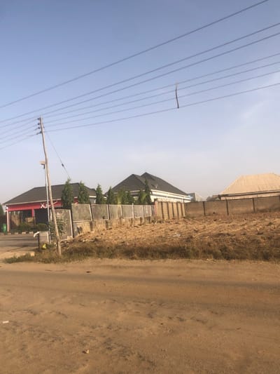 A street view showcases a dirt road alongside a building with a dark roof and a row of solar panels. Another building is visible in the background. Above, a power pole supports a series of electrical wires. A stick is precariously perched on one of the wires. The sky is pale blue.