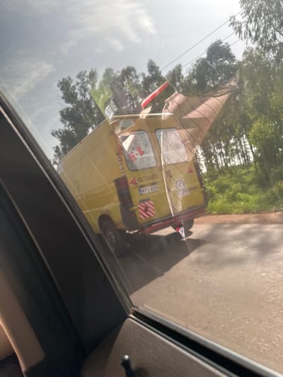 A yellow van with "IOLTOAUTO" branding is seen from a side window, possibly Kassa Market, Nigeria, on a road. The van is decorated with red and white striped tape on the rear bumper and has a license plate that reads "KVI-825." There are flags attached to the roof, one with red and white stripes. Trees and power lines are visible in the background.