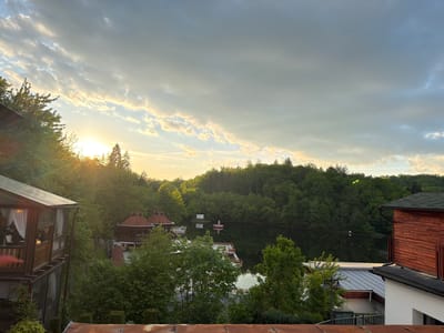 The image captures a scenic view in Sovata, Romania, featuring a lake surrounded by lush green trees under a partly cloudy sky at sunset. In the foreground, the image includes rooftops and the architecture of nearby buildings, including a wooden building with a covered porch. On the left, a multi-story structure has glass enclosed sections with white curtains. Across the water, several wooden gazebo-like structures sit on docks, reflected in the still water of the lake. A small white structure floats in the lake, closer to the dense treeline, which slopes gently up to meet the sky. The setting sun casts a warm glow through the trees on the left side of the image.
