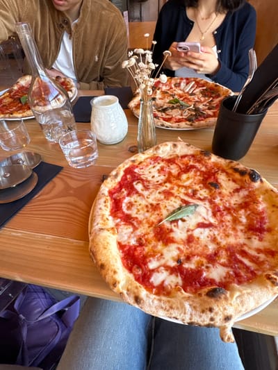 The image captures a top-down, slightly angled view of a dining table, rotated ninety degrees counter-clockwise. The scene shows two pizzas and several other items on a light wooden table, with parts of two individuals visible.
In the foreground, dominating the right side of the frame, is a large, round Neapolitan-style pizza. It features a prominent, puffed crust with visible charring, indicating a high-temperature bake. The pizza is topped with a vibrant red tomato sauce and melted white mozzarella cheese, which has a bubbly texture. A single green leaf, likely basil or sage, is placed near the center of the pizza.
Towards the middle and left side of the table, another smaller pizza is visible, partially obscured by other items. This pizza appears to have toppings like mushrooms and possibly other vegetables or herbs, differing from the simpler Margherita-style pizza in the foreground.
Several dining accessories are scattered across the table. A tall, black cylindrical holder filled with various cutlery (forks and knives) is positioned towards the top left. Below this, a slender glass vase holds a small bunch of dried white, spherical flowers or grass. A small, white, textured ceramic jug or vase is also on the table.
Numerous clear glass items are present: several empty drinking glasses are arranged, some upright and some tilted. An empty glass bottle, shaped like a wine bottle but possibly decorative, lies on its side, reflecting light. A silver pizza cutter with a black handle rests on a black placemat.
Two individuals are partially visible at the table. In the upper left corner, a person with dark hair is seen wearing a dark blue cardigan with light buttons over a white top. They are holding a light pink smartphone, seemingly taking a picture or video of the food. A delicate gold necklace with a circular pendant is visible around their neck. In the lower left, another person is partially shown, wearing a brown corduroy jacket over a white top. Only their torso and arm are visible, and they appear to be looking down at the table.
To the far right of the image, the leg of a person wearing blue jeans is visible, suggesting someone is seated at the table on a bench or chair. In the very bottom right corner, a purple bag, possibly a backpack or tote, is partially visible on the floor or a nearby seat.
The overall impression is that of a casual meal at a restaurant or cafe, with people enjoying food and one person capturing the moment with their phone. The lighting suggests daytime.