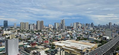 This is a high-angle, wide shot of Makati City, Philippines, on a cloudy day. The city is densely populated with a mix of residential and commercial buildings, including numerous high-rise apartment buildings and office towers. A multi-lane elevated highway with light traffic curves through the foreground. The buildings vary in architectural style and age, with some appearing modern and sleek, while others are older and more traditional. Greenery is visible between some of the structures, suggesting parks or street trees. The sky is filled with a layer of cumulus clouds, obscuring the sun and creating a diffused light. The overall impression is one of a sprawling, active urban environment.