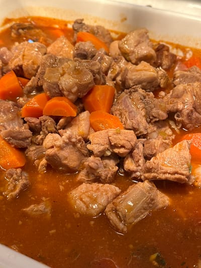 This image depicts a close-up, overhead view of a cooked stew or braise served in a white, rectangular dish. The scene is located in Púng Luông, Vietnam. The dish primarily contains numerous irregular chunks of light brown meat, consistent with pork, interspersed with bright orange, uniformly diced carrots. All ingredients are coated and partially submerged in a thick, reddish-orange sauce that exhibits a glossy surface. Small, indistinct flecks, possibly herbs, spices, or finely minced vegetables, are visible within the sauce. The meat pieces show varying textures, with some appearing lean and others presenting visible fatty or connective tissue. The carrots appear softened from cooking. The white serving dish has slightly angled edges. The background outside the immediate dish is out of focus and indistinct. No people, specific actions, or external environmental details beyond the serving dish are discernible.