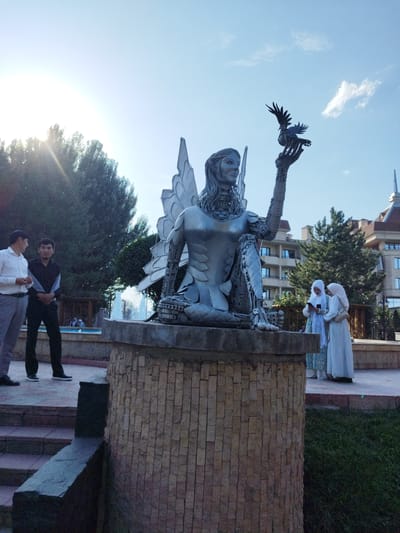 The photograph captures a statue of a winged figure holding a bird, set in an outdoor public space. The statue, made of metal, depicts a female figure with elaborate wings and intricate detailing on her body. She is kneeling and extending her hand, on which a bird perches with its wings spread. The statue stands on a cylindrical pedestal covered with light-colored stone tiles.
In the foreground, two men are standing on a paved area. One is wearing a white dress shirt and light-colored trousers, while the other is in a black shirt and black trousers. Further back, two women wearing hijabs are looking at their phones. Behind the statue, a fountain sprays water, and in the background, there are trees, buildings with arched windows, and a clear blue sky with a few scattered clouds. The sun is shining brightly, casting strong shadows, indicating it is likely daytime. The overall scene appears to be a park or a public square in Kenbulun, Kyrgyzstan.
