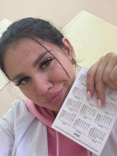 A young woman with dark hair, green eyes, and prominent false eyelashes poses for a selfie, holding a portion of a 2026 calendar in her right hand. She is wearing a pink hooded sweatshirt under a white jacket. The calendar displays months in Russian, including "Январь" (January), "февраль" (February), "Март" (March), "Апрель" (April), "Май" (May), "Июнь" (June), "Июль" (July), "Август" (August), "Сентябрь" (September), "Октябрь" (October), "Ноябрь" (November), and "Декабрь" (December). The text at the bottom of the calendar reads "ИДЕАЛЬНОЙ ПРИЧЁСКОЙ ДЛЯ ГРУДНОГО РЕБЁНКА ЯВЛЯЕТСЯ МОЛОКО МАТЕРИ" which translates to "The ideal hairstyle for a baby is mother's milk". The background appears to be an indoor setting with a cream-colored textured surface and a wall. No specific location in the city of Гаревая, Russia is discernible from the image.