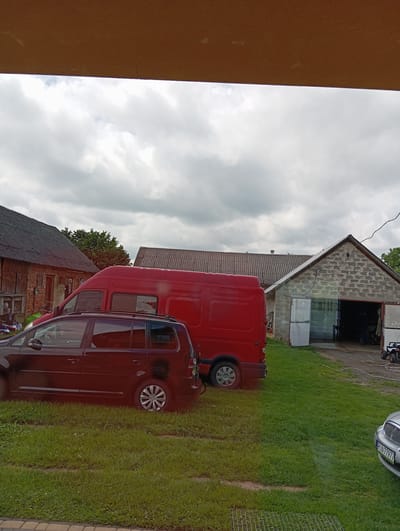 The image depicts an outdoor scene in Hlynytsi, Ukraine. In the foreground, a dark-colored station wagon is parked on a grassy area. Behind it, a bright red van is also parked. To the right, a concrete building with a corrugated roof and two open garage doors is visible. Inside the garage, some objects and a person can be partially seen. Further to the left, a brick building with a dark, corrugated roof is present. The sky above is overcast with clouds. The ground is covered with green grass.