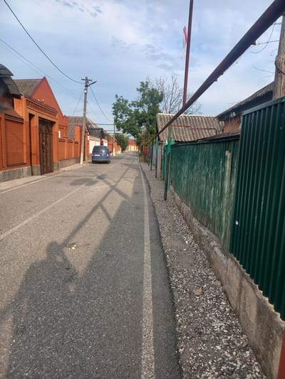 The image depicts a street scene in Argun, Russia, with buildings on either side and a single blue car parked on the asphalt road. The buildings feature red brick facades and corrugated metal roofs. A tall, dark green corrugated metal fence runs along the right side of the street, separating the road from the properties behind it. The road is paved with asphalt and has white lane markings. In the foreground, the shadow of a person holding a camera is cast on the road, indicating the photographer's presence. The sky is partly cloudy, suggesting daytime. The environment appears to be a residential area with a somewhat weathered appearance, characterized by peeling paint on the fence and gravel along the roadside. A flagpole with a tattered flag is visible in the distance.