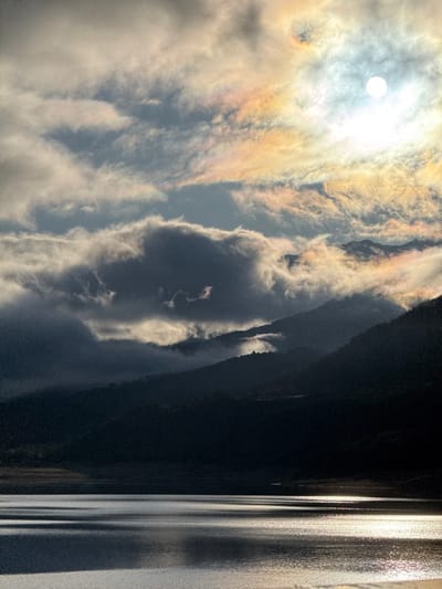 The image presents a natural landscape within the vicinity of Savines-le-Lac, France, likely featuring Lac de Serre-Ponçon. The foreground and lower mid-ground are occupied by a body of water exhibiting horizontal reflections of sunlight, primarily on its right side. Dark, undulating mountain ranges and hills rise from the water's edge, appearing silhouetted against the sky due to backlighting. These elevations are partially obscured by layers of low-hanging clouds and fog, which vary in density and illumination. The sky dominates the upper half of the frame, featuring a distinct, brilliant white sun located in the upper right quadrant. This sun is surrounded by clouds exhibiting iridescence, displaying hues of yellow, orange, pink, and light blue. Other clouds across the sky range from bright white to deep grey. No individuals, specific man-made structures, or human activities are visible.