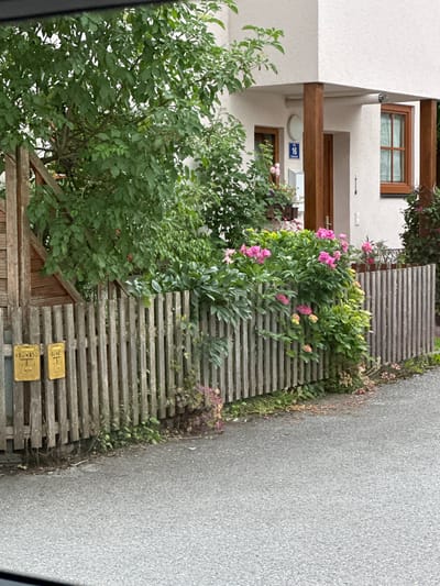 The image depicts a residential property in Walserfeld, Austria, featuring a wooden fence bordering a paved road. Behind the fence, lush green foliage and pink flowering plants are visible, adjacent to a modern-style house with a white exterior and brown window frames. A blue house number sign with the number "16" is affixed to the house. Two yellow rectangular signs with black text are attached to the fence. The environment appears to be a well-maintained outdoor area, likely a garden or front yard. No people are present in the image, and no actions are being depicted.