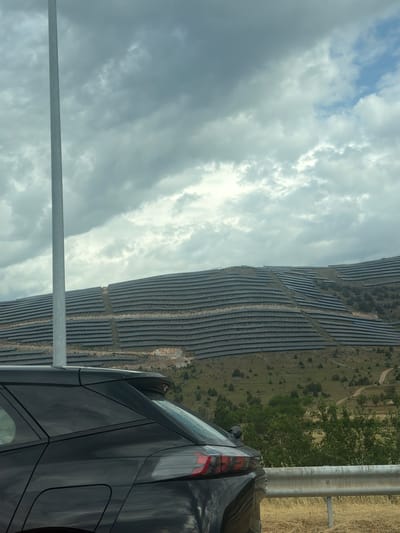 The image shows a large solar panel farm situated on a terraced hillside. The solar panels are dark and arranged in neat rows that follow the contours of the terrain. The hillside is covered with sparse vegetation, including some scattered trees and dry grass. In the foreground, a portion of a black car is visible, with its rear end and roof showing. A metallic guardrail runs horizontally across the lower right section of the frame. A tall, slender, light-colored pole stands vertically on the left side of the image, extending upwards into a cloudy sky. The sky itself is filled with a mix of dark, heavy clouds and patches of brighter, lighter clouds, with a hint of blue peeking through in the upper right corner. The scene is likely an outdoor environment, possibly a rural or semi-rural area, given the presence of the solar farm and the natural landscape. The location is specified as Αλωνάκια, Greece.