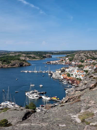 The image depicts a panoramic view of the picturesque coastal town of Fjällbacka, Sweden. A vibrant blue sky with a few wispy clouds stretches across the top of the frame. Below, the deep blue water of a harbor is dotted with numerous boats of various sizes, including sailboats with their masts raised, sleek motor yachts, and smaller dinghies. The boats are moored at piers and anchored in the water, creating a lively maritime scene.
The town itself is nestled on the rocky shoreline, with a cluster of charming houses characterized by their red-tiled roofs and white or light-colored facades. The architecture suggests a traditional Scandinavian coastal village. The buildings are tightly packed, cascading down the slopes of the rugged, greyish-brown rocks that dominate the landscape. These rocks are partially covered with sparse green vegetation, adding a touch of natural beauty to the scene.
In the background, the coastline continues with more rocky outcrops and patches of dense green forest. The overall impression is one of a serene and idyllic seaside community, bathed in bright sunlight on a clear day. The perspective suggests the photo was taken from an elevated vantage point, likely a hillside overlooking the harbor and town. There are no visible people in the immediate foreground or distinct actions, but the presence of numerous boats implies a bustling activity within the harbor.