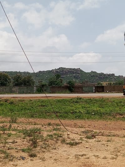 The image depicts a landscape scene in Unguwar Saya, Nigeria. The foreground is dominated by dry, sandy earth with sparse patches of green grass and vegetation. A single wire runs diagonally from the top left to the bottom right of the frame, appearing to be a power line or telephone wire.
In the middle ground, a paved road runs horizontally across the image, bordered by a low, greenish wall constructed of concrete and brick. Beyond the wall, a dense area of trees and foliage can be seen, with some buildings partially visible.
The background features a prominent rocky hill or mountain range, covered in green vegetation and dotted with large boulders. The sky above is partly cloudy with a light blue hue, suggesting daytime. The overall impression is one of a rural or semi-rural environment with natural and man-made elements coexisting. No people are visible in the image, and the activity or event taking place is not evident. The weather appears to be fair. No discernible text is present in the image.