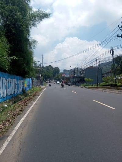 A street scene shows an asphalt road with a yellow line dividing traffic lanes, bordered on the left by a white line and a blue wall with graffiti, possibly saying "BIDONG 1933". Trees and vegetation are on the left side. Several motorcycles are on the road, heading in the opposite direction, along with a white vehicle farther away. Buildings and utility poles with many wires are visible on the right side of the road, with a hillside in the background. The sky is partly cloudy, with a mix of blue sky and large white clouds. The location is likely in an urban or suburban area. It's not possible to confirm the location is Nawng Hkar, Myanmar, without further information.
