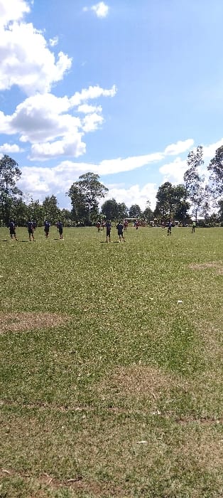 The image shows a grassy field with approximately 20 individuals in the distance. The individuals are spread out across the field, and some appear to be engaged in a game. The sky is blue with white clouds. Trees line the perimeter of the field. The location is a field in Bungoma, Kenya, with the coordinates 0.554 latitude, 34.774 longitude. The location is likely in a rural area. The grass is green with some brown patches, suggesting the field is not perfectly manicured. The image was likely captured on a sunny day, based on the clear blue sky and the shadows cast by the individuals.