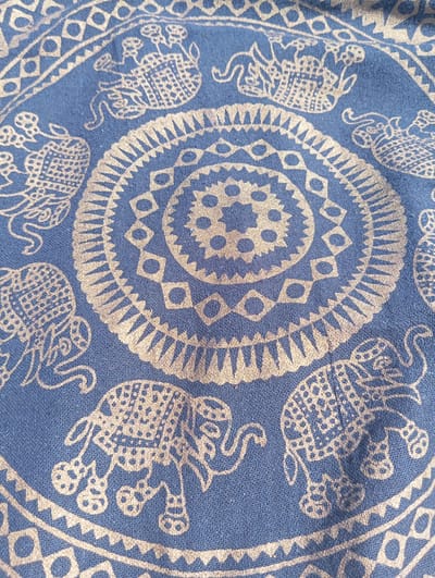 The image shows a close-up of a blue fabric with a white or light gold patterned print. The pattern is circular and features repeating motifs of elephants and geometric shapes, including circles and diamonds. The fabric appears to be a textile, possibly a scarf or tapestry, with a visible weave texture. The lighting suggests the fabric is illuminated from the top right, casting subtle shadows. There are no people or specific locations in Wasilków, Poland visible in the image.