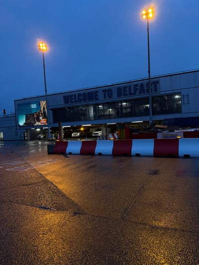 This image displays the exterior of Belfast International Airport, located in Crumlin, United Kingdom, during twilight or night conditions. The sky is dark blue, and the asphalt pavement is wet, reflecting the artificial lights.
The primary subject is the airport terminal building, which features a prominent sign across its facade stating "WELCOME TO BELFAST" in large blue lettering. To the left of this sign, a large billboard displays an advertisement for "BRILLIANT CITY BREAKS AT UNBEATABLE PRICES" by "Jet2 Holidays," alongside an image of an ancient-looking stone archway. The ground level of the building includes a brightly lit, covered drive-through area where at least three light-colored vehicles, including one SUV, are visible. A person wearing a high-visibility vest is partially discernible near the vehicles on the left side of this area.
Two tall, grey metallic light poles, each topped with four amber floodlights, stand in the foreground and midground, casting strong illumination onto the wet pavement. A continuous line of red and white plastic traffic barriers separates the foreground area from the building's immediate entrance. Behind these barriers, a dark blue or black tarpaulin covers a low structure, and a yellow sign with the word "MINSTER" is visible on the right. The wet pavement exhibits reflections of the lights, with faint surface cracks and subtle white markings.