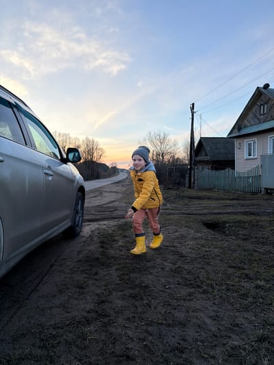 A photograph taken in Дулесово, Russia, shows a young child in a yellow jacket, brown pants, and yellow boots. They also wear a grey beanie. The child is on a dirt road beside a grey car. The background includes a house, a telephone pole, bare trees, and a sky with soft, diffused light.