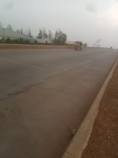 The image depicts a hazy, overcast day in Sabon Gari, Nassarawa, Nigeria. The foreground is dominated by a wide, paved road, likely a highway or major thoroughfare, with a distinct curb on the right side separating the road from a reddish-brown dirt embankment. The road surface appears relatively new, with some tire marks visible. In the mid-ground, a yellow three-wheeled vehicle, possibly a tuk-tuk or auto-rickshaw, is traveling on the road. Further in the background, buildings with green roofs are visible, along with sparse vegetation and trees. The overall atmosphere is subdued due to the overcast conditions.