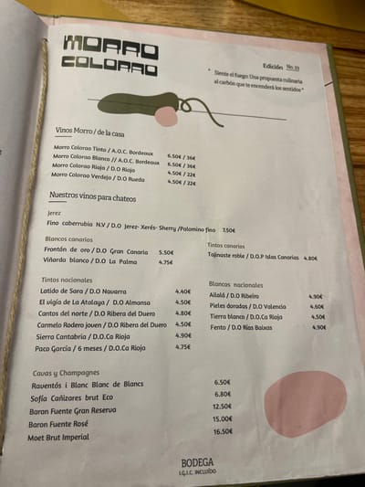 A menu from a restaurant named "Morro Colorado" is presented. It is printed on a white paper, and the text is black, with some prices highlighted in red. The menu lists various wines, including whites, reds, and sparkling wines. The wines are categorized by origin and type. Some wines are labeled as "our wines for tapas." An image of a red, white, and black heart-shaped design is in the top left corner. The menu is from the restaurant Morro Colorado, located in Las Palmas de Gran Canaria, Spain (28.102, -15.414).