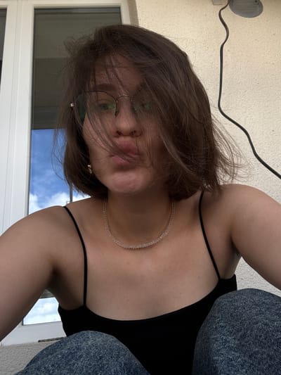 A young person with short, brown hair and glasses is taking a selfie outdoors. They are wearing a black tank top and blue jeans. Their face is partially obscured by their hair and they are puckering their lips. A clear beaded necklace is visible around their neck. The background shows a textured wall and a window with a view of the sky. There is a black cord running along the wall. There is no indication of the city of Pervomaysky, Russia in the image.
