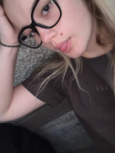 The image shows a close-up of a young person wearing black-rimmed glasses and a dark brown t-shirt with the embroidered text "ELS." Their left hand is resting on their head, and their tongue is sticking out. The background appears to be a textured, grey surface. The image is a selfie, taken from a slightly upward angle. There are no discernible landmarks or indicators that would suggest the location is specifically in Karlovo, Bulgaria.
