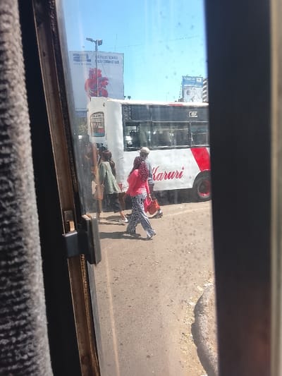 The image captures a street scene in what appears to be a bustling urban or semi-urban area, seen from inside a vehicle through a window. The window frame is visible on the left and right, and specks on the glass suggest dirt or reflections. Bright daylight and a clear blue sky indicate it is daytime, likely mid-day.
In the foreground, several pedestrians are walking on a paved surface next to a large white public transport bus. A woman in a pink top and black-and-white striped pants walks towards the right, carrying a red bag. Next to her, a man in a light-colored cap and dark clothing also walks, carrying a brown bag. Other people are visible near the bus, some potentially boarding or disembarking.
The prominent white bus features a red diagonal stripe and the word "Karuri" written in a cursive, red font on its side. Part of the word "THE" is visible above the red stripe near the back of the bus. In the background, above the bus, two large billboards are visible. One prominent billboard advertises "AL wire & cable" with the slogan "THE PULSE BEHIND EVERY CONNECTION" and features an abstract, red, heart-like graphic of interconnected wires. Further in the background, another billboard shows partially visible text, including "Dr. Agarwal's".
The scene depicts everyday activity on a street, with pedestrians and a public bus. The presence of the bus named "Karuri" provides a strong location cue, aligning with the context of Kiambu Township ward, Kenya, where Karuri is a known area. The overall impression is one of a busy, sunny day in a Kenyan urban landscape.