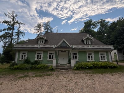 The image shows the exterior of a traditional wooden house, painted in a pale green color, in Bialowieza, Poland. The house has a dark-colored, shingled roof with several dormer windows and decorative white trim along the eaves and around the windows. The front of the house features a central entrance with a double white door, reached by a set of stone steps. There are multiple white-framed windows on the facade, some adorned with intricate white wooden carvings. Lush green ferns and other foliage grow along the base of the house, and tall pine trees are visible to the left. The foreground is a gravel path, with a cobblestone section closer to the house. The sky above is blue with scattered white clouds. The scene appears to be set in a natural, possibly wooded, environment. There are no people or animals visible in the image.