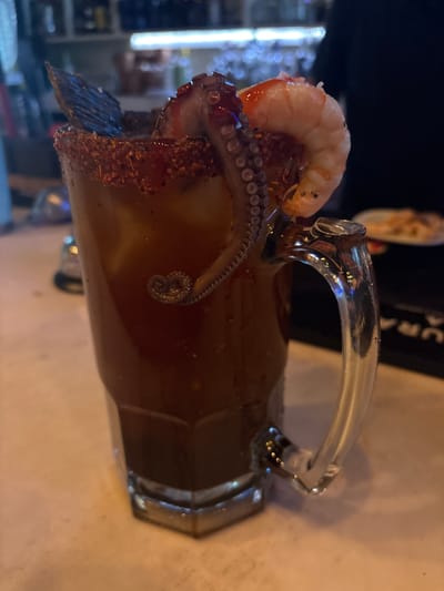 A large glass mug filled with a dark liquid stands on a light-colored surface. The rim of the mug is garnished with a reddish-brown substance. The mug is adorned with seafood: a cooked shrimp perched on top and a large octopus tentacle dangling over the side, with some of the tentacle submerged in the liquid. The octopus tentacle has visible suckers. A dark, possibly smoked, fish piece is also attached to the rim. The mug has a clear glass handle. The background shows a blurred cityscape with bright lights, suggesting an urban setting. This photograph appears to be taken in Santiago de Querétaro, Mexico.