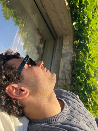 A man with curly dark hair is reclined, wearing black Ray-Ban sunglasses and a grey cable-knit sweater. He is looking up towards a green, overgrown structure that appears to be a rooftop garden. The sunlight reflects off a window, mirroring the greenery and sky. The location is consistent with Gondarém, Portugal, a coastal city known for its greenery and modern architecture often incorporating rooftop gardens.