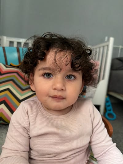 A baby with curly dark hair and blue eyes is looking up at the camera. The baby is wearing a long-sleeved pink shirt. The baby is lying on a surface with a colorful zig-zag pattern. Behind the baby, there is a white crib, a gray wall, and a blue toy. The location in Test Valley, United Kingdom, cannot be determined from the image.