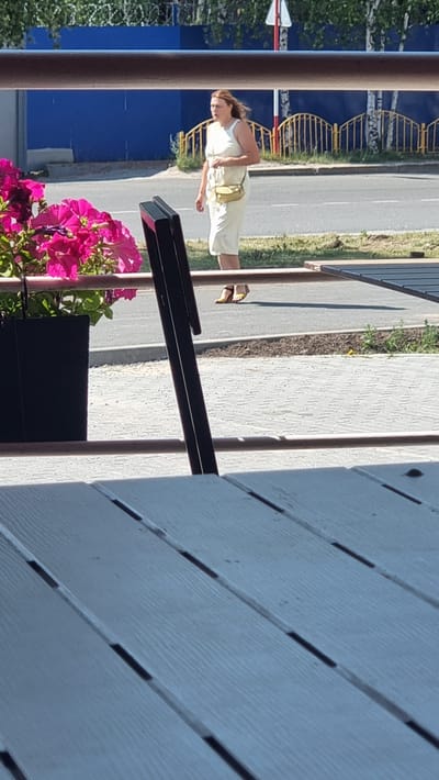 A woman is walking near a street in the city of Megion, Russia. She is wearing a light-colored dress and carrying a yellow purse. The woman is walking on a sidewalk. In the foreground, there is a table and some flowers in a pot. In the background, there is a blue wall and a yellow fence. The weather is sunny.