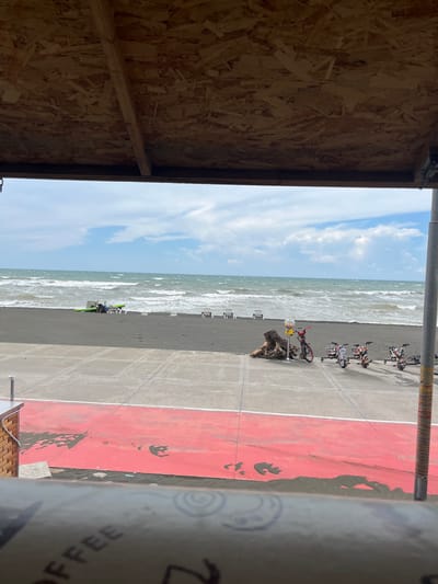 This media file shows a scene at a beach in Ureki, Georgia. The perspective is from inside a wooden structure, looking out towards the Black Sea.

The foreground of the image is dominated by the interior of a wooden structure with visible particle board walls and wooden beams. Outside, the scene unfolds with a sandy beach, the sea, and a paved area with red markings. Several bicycles are parked on the paved area, some with colorful decorative elements. A small gumball machine sits atop a piece of driftwood on the beach. Further out on the beach, lounge chairs are visible, as well as some beach equipment, possibly including a green kayak or paddleboard, near the water's edge. The sea is moderately rough with visible waves and white foam. The sky is partly cloudy with blue patches.

The time of day appears to be daytime, likely late morning or early afternoon, given the brightness of the scene and the angle of the sunlight, though this is not explicitly clear. The weather seems to be warm and possibly breezy, as indicated by the choppy sea. The presence of beach furniture and recreational equipment suggests a typical day at a beach resort.

There is visible text on a cylindrical object in the right-hand portion of the image, which appears to be a paper cup. The visible words are "COFFEE" and some unreadable text below it.

The overall impression is one of a recreational beach setting, potentially on a holiday or a leisurely day. There are no people prominently visible in the frame, adding to a sense of calm or a pause in activity.
