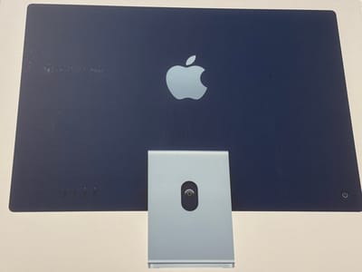 The image shows the back of a dark blue Apple iMac computer. The Apple logo is prominently displayed in the center of the screen. At the bottom of the screen, there are four small indicators, possibly for status lights, and a power button icon in the lower right corner. The computer is resting on a light-colored surface, and its silver stand is visible at the bottom. The surface appears to be a desk or table. There are some light smudges and scratches on the back of the iMac. No people or specific activities are depicted in the image. The setting is indoors, and there are no indications of the time of day or weather.