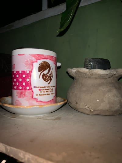 A close-up, eye-level shot shows a pink and white patterned mug sitting on a saucer on a light-colored, textured surface. The mug has a decorative label with text and a silhouette of a woman's profile with flowers. To the right of the mug, there is a rough, light brown, pot-like object with a dark object placed on top of it. The background is a dark green wall with a narrow white ledge above it, and a green leaf is visible hanging from the top edge of the frame. The lighting appears to be artificial, creating shadows and highlighting the textures of the objects. The scene is indoors or in a sheltered outdoor space, likely at night given the darkness. Based on the text on the mug, which includes "Kestugihan Kidul - Cilacap", this scene is likely located in Cilacap, Indonesia, possibly Karangkemiri, Indonesia, given the provided context.