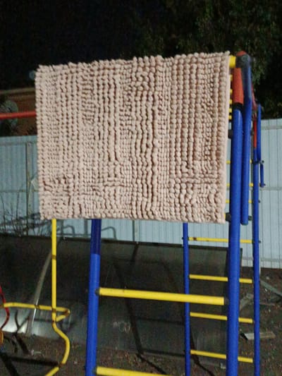 A beige mat is draped over a section of a blue and yellow playground structure. The mat is textured with many small, raised loops. The playground structure appears to be made of metal pipes. In the background, a light-colored metal fence is visible, and beyond that, dark trees. The ground beneath the structure is covered with debris and dry leaves. The scene is dimly lit, suggesting it is nighttime or dusk. There are no people visible in the image. This scene is located in the city of Novotitarovskaya, Russia.