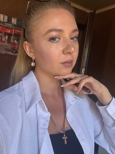 The image shows a close-up of a young woman with blonde hair pulled back, wearing a white collared shirt and a gold cross necklace. She has on earrings, rings, and makeup. Her hand is resting on her chin, and she is holding a cylindrical object, possibly an e-cigarette, in her hand. In the background, there is a wall with a framed picture of a cafe with a red facade and a red scooter. The overall scene is indoors, with a wooden closet door visible on the right. The location is stated to be in Makiivka, Ukraine.