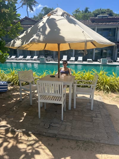 A woman is seated at a white wooden table and chairs next to a swimming pool. She is wearing a purple bikini and sunglasses. The table is set with a dark object, possibly a phone. Behind the table, lush green and yellow plants border the turquoise water of the pool. In the background, a hotel building with multiple balconies and windows is visible. A large cream-colored umbrella shades the table and chairs, casting dappled shadows on the paved walkway. The setting appears to be a resort or hotel in Mirissa, Sri Lanka, during the daytime, indicated by the bright sunlight and clear blue sky. The overall mood is relaxed and leisurely, suggesting a vacation or a break by the pool. There is a visible "SOLARAY" logo on a solar water heater attached to the hotel building.