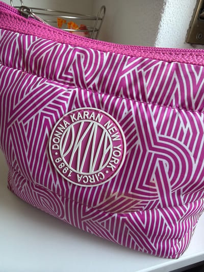 A close-up, slightly angled shot shows a pink and white geometric patterned toiletry bag, likely made of nylon, resting on a white surface. The bag features a textured pink band along the top edge, with a silver zipper partially visible. A circular emblem with the text "DONNA KARAN NEW YORK CIRCA 1989" in white is prominently displayed on the front of the bag. The background is out of focus but appears to be a light-colored, textured wall. There are no people or animals in the image. The scene is indoors, on a clean white surface. There is no indication of a location in Rupperswil, Switzerland.