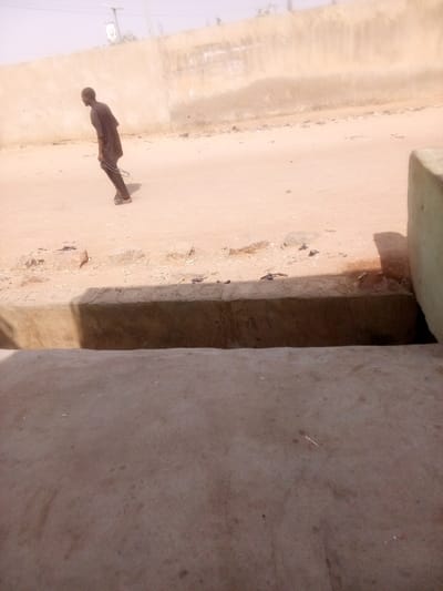 The scene depicts an outdoor environment in Tudun Murtala, Nigeria. The foreground consists of a grey concrete surface, followed by a lower concrete ledge that casts a dark shadow. To the far right, a partial light-green painted concrete block structure is visible. Beyond the ledge, an unpaved, reddish-brown dirt path stretches diagonally, scattered with small rocks and dark debris.
A single individual, appearing male, is present on this path, walking from left to right. He is dressed in dark clothing, consistent with a tunic and trousers, and wears sandals. He holds a dark, chain-like object in his right hand, which extends downwards near his left leg.
The background features a tall, light-brown textured wall or barrier, extending across the frame. Above the wall, a hazy sky is visible, with a faint outline of what appears to be a utility pole or antenna on the far left. Strong sunlight from the upper right illuminates the scene, casting distinct shadows.