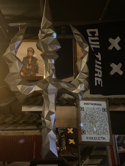 The image depicts an installation in Thong Sala, Thailand, featuring a metallic sculpture resembling a stylized skull or mask. A banner with the word "CULTURE" in white block letters is visible in the background, adorned with two stylized "X" marks. A QR code labeled "INSTAGRAM @XX.CLOTH" is affixed to a wooden structure adjacent to the banner. The sculpture frames a partial view of a person wearing patterned clothing and headwear. The environment appears to be a dimly lit, possibly urban or commercial space.
