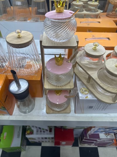 The image displays a retail environment, likely a market stall or shop shelf, exhibiting a collection of kitchenware. The focus is on glass jars with wooden lids and gold-colored crowns, some containing pink lids with spoons. These are arranged on wooden tiered stands and a wooden tray. Other items visible include more glass jars with similar wooden lids, some appearing to be part of a set, and a glass dispenser with a metal spout. In the background and lower sections of the image, various boxed items, including what appear to be tissue boxes and other packaging, are present. The overall color palette is dominated by clear glass, wood tones, and hints of pink and gold. The setting appears to be indoors, on shelving, with a checkered floor visible at the bottom. The scene is static and shows no people or active interactions. The location is specified as Kara-Suu, Kyrgyzstan.